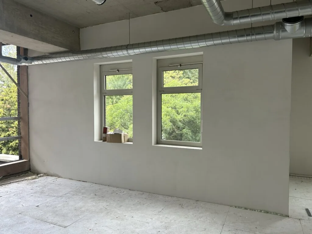 Binnenruimte in verbouwing met twee ramen, zicht op groen buiten en zichtbare ventilatiebuizen aan het plafond.